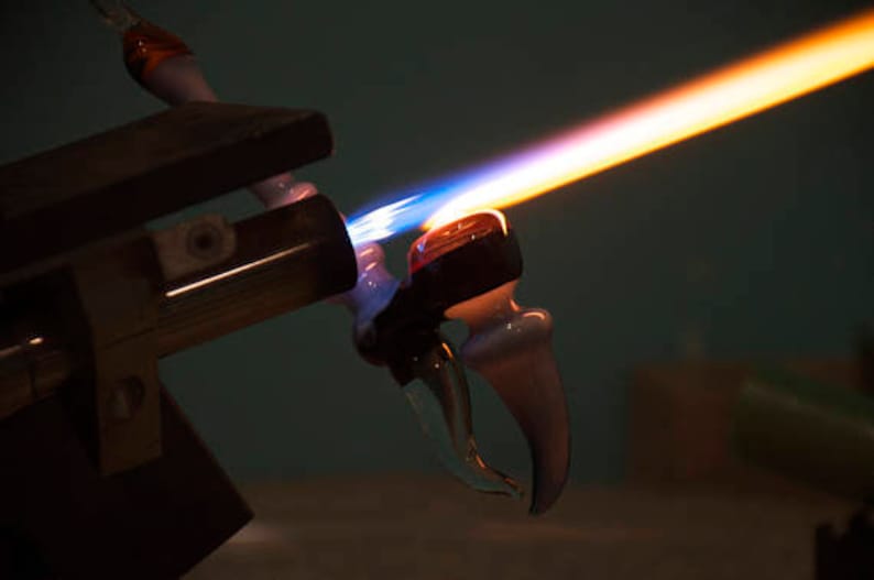 Op de afbeelding: Een glasblaasbrander met een blauwe en oranje vlam verhit een glazen pijp. De pijp wordt gevormd tot een gebogen ontwerp.