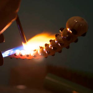 Op de afbeelding: Een glazen pijp die met een brander wordt verwarmd. De pijp is gevormd als een rups met zwarte stippen en een lichtbruin lichaam.
