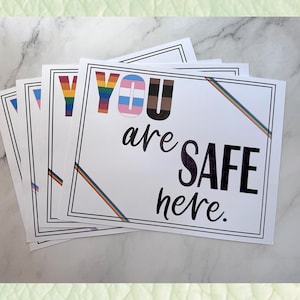 Puede incluir: Una pila de letreros rectangulares blancos con bordes negros. Los letreros presentan el texto "YOU are SAFE here". La palabra "YOU" está en colores del arcoíris, y la palabra "SAFE" es morada. Los letreros tienen rayas de arcoíris.