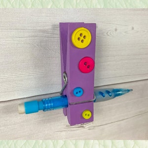 May include: Purple wooden clothespin with four colorful buttons and a blue pencil held in place by the clothespin.