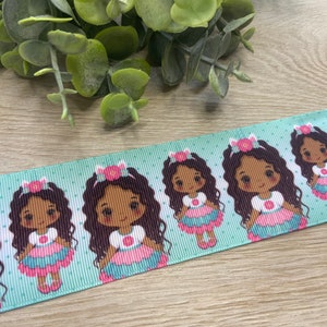 May include: A light blue ribbon featuring a repeating pattern of a cartoon girl with dark hair, wearing a pink and blue dress and a flower crown. The ribbon has a polka dot design.