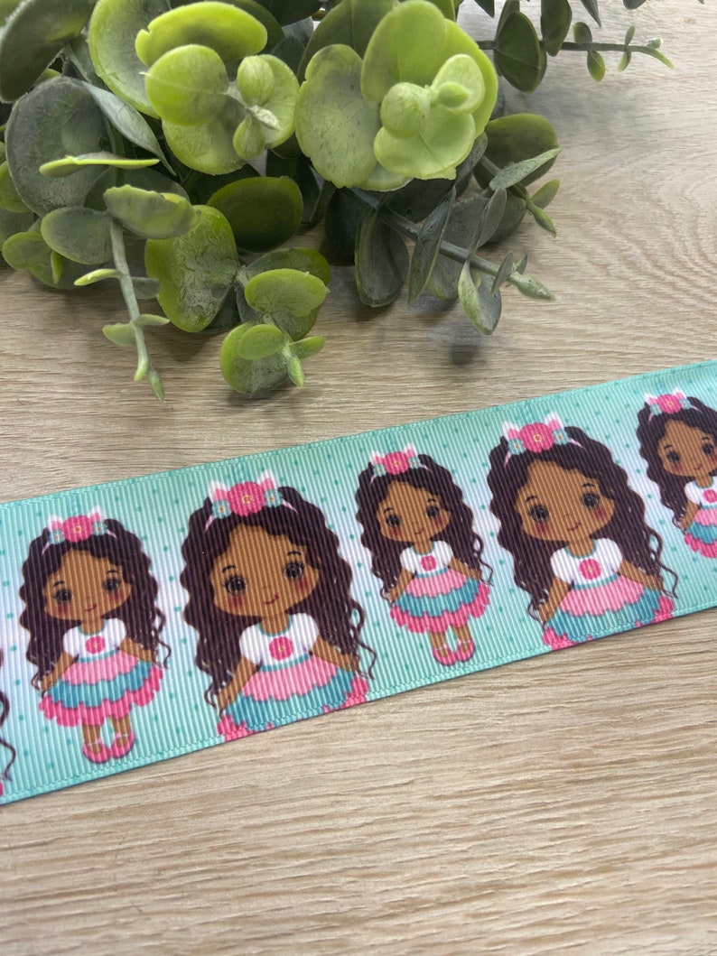 May include: A light blue ribbon featuring a repeating pattern of a cartoon girl with dark hair, wearing a pink and blue dress and a flower crown. The ribbon has a polka dot design.