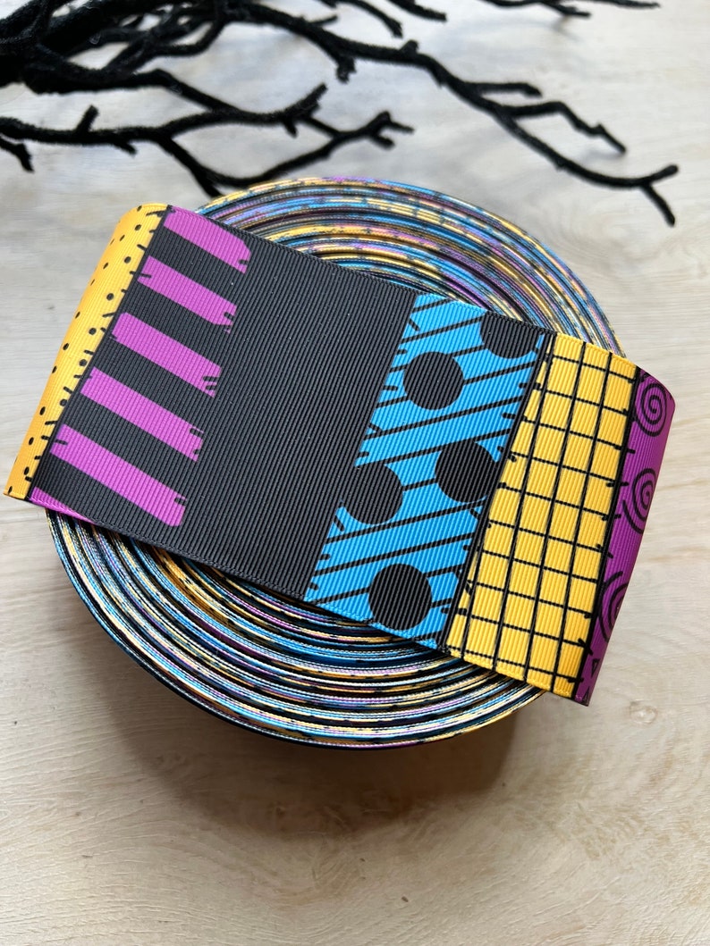 May include: A roll of patterned ribbon featuring a patchwork design in black, yellow, purple, and blue. The ribbon has a variety of patterns, including stripes, polka dots, and a grid. The ribbon is sitting on a roll of iridescent ribbon.