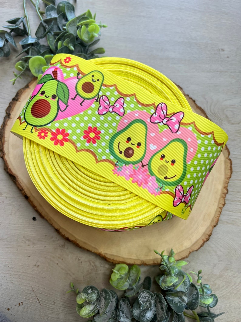 May include: Yellow ribbon with a repeating pattern of green avocados with pink bows and white polka dots. The avocados have cute faces and are holding hands.