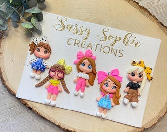 May include: Six colorful polymer clay doll charms with different hairstyles and outfits. The dolls are wearing pink, blue, yellow, and brown clothing. The dolls have brown, blonde, and black hair. The charms are on a white background with the text "Sassy Sophie Creations".