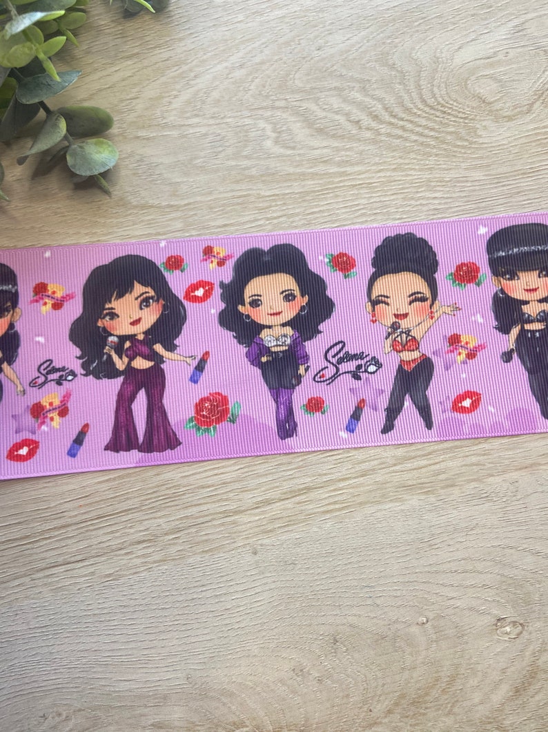 May include: Purple ribbon with a repeating pattern of cartoon figures of women with red roses and lipstick. The text "Selena" is printed on the ribbon.
