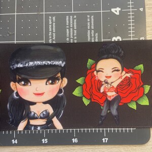 Pu&ograve; includere: Nastro bianco e nero con un motivo ripetuto di una donna dei cartoni animati che indossa un cappello nero e un abito nero e argento, e una rosa rossa con foglie verdi.