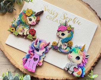 May include: Four colorful wooden unicorn ornaments with glitter accents and floral details. The unicorns are all different colors and have different expressions. The ornaments are on a wooden background with the text "Sassy Sophie Creations".