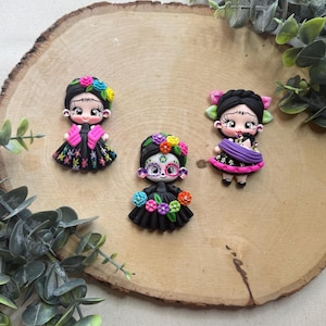 May include: Three colorful handmade polymer clay figurines of women wearing traditional Mexican clothing. The figurines are on a wooden surface.