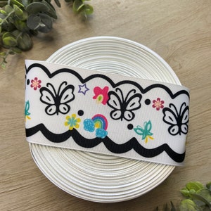 May include: A white ribbon with a black scalloped edge and a colorful print of butterflies, flowers, and a rainbow. The ribbon is rolled up and ready to be used for crafting or decorating.