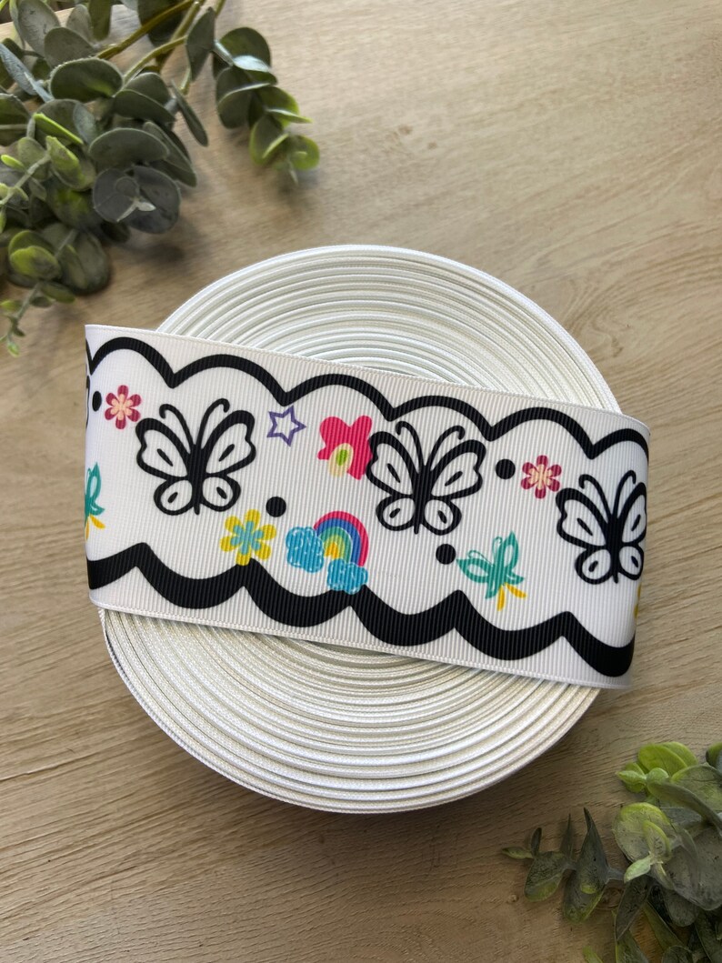 May include: A white ribbon with a black scalloped edge and a colorful print of butterflies, flowers, and a rainbow. The ribbon is rolled up and ready to be used for crafting or decorating.