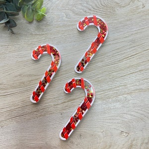 May include: Three candy cane-shaped ornaments with red and white stripes and filled with red and gold glitter. The ornaments have a clear, see-through center and a white border. The candy canes are arranged on a light wood-grain surface.