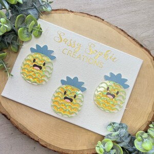 May include: Three yellow and green glitter pineapple resin ornaments with blue tops and happy faces. The ornaments are on a white canvas with the text "Sassy Sophie Creations" in gold lettering.