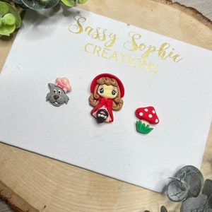 May include: A set of three red, white, and gray clay charms featuring a girl in a red hooded cloak, a wolf wearing a pink hat, and a red and white mushroom with green grass.