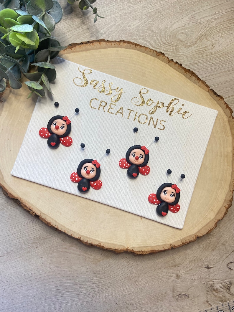 May include: Four black and red ladybug ornaments with white polka dots and black eyes. The ornaments are displayed on a white canvas with the text "Sassy Sophie Creations" in gold glitter lettering.
