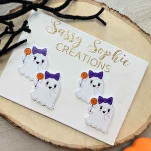 May include: Four white ghost-shaped Halloween decorations with purple bows and orange lollipops are displayed on a white canvas. The canvas has the text "Sassy Sophie Creations" in gold glitter. The canvas is on a wooden slice.