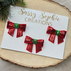 May include: Three red sequined bows with green centers are displayed on a white canvas. The canvas has gold text that reads "Sassy Sophie CREATIONS". The canvas is placed on a wooden surface, with a sprig of greenery and red berries in the upper left corner.