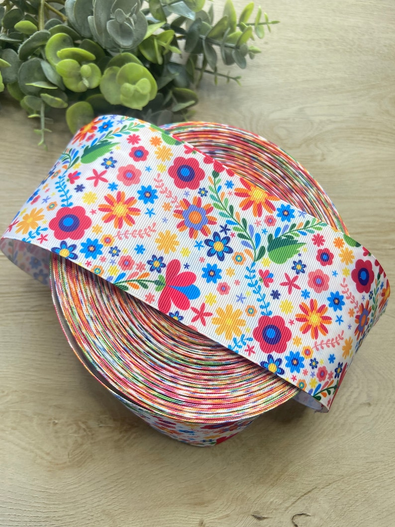 Puede incluir: Un rollo de cinta blanca con un estampado floral colorido. La cinta presenta flores rojas, amarillas, azules y verdes sobre un fondo blanco.