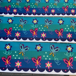 Pu&ograve; includere: Un nastro blu e verde con un bordo bianco a festoni. Il nastro presenta un motivo ripetitivo di farfalle e fiori. Le farfalle sono rosa, gialle e blu. I fiori sono bianchi con centri rosa.
