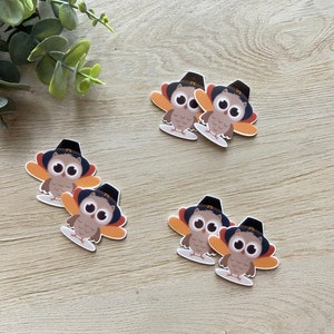 May include: Several owl-turkey hybrid stickers are arranged on a light wood surface. Each sticker features a cartoon owl with a pilgrim hat and turkey-like wings. The owls are brown, with orange and white accents.