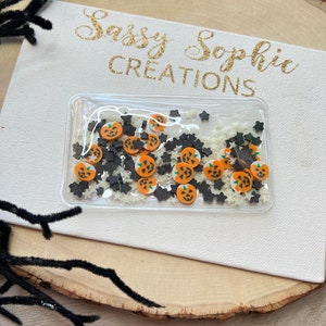 May include: A clear rectangular container filled with orange pumpkin and black star-shaped confetti. The container sits on a white canvas with gold text that reads "Sassy Sophie Creations". A black, fuzzy branch is in the upper left corner.