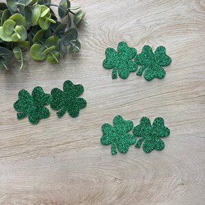 May include: Four green glitter shamrock cutouts on a light wood background. The shamrocks are arranged in two groups of two.