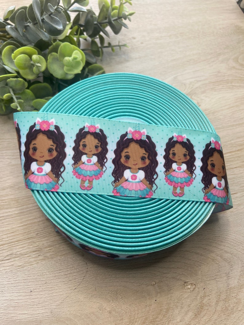 May include: A roll of turquoise ribbon with a repeating pattern of cartoon girls in dresses. The ribbon is approximately 1 inch wide and is suitable for crafts, gift wrapping, or hair accessories.