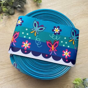 Pu&ograve; includere: Un rotolo di nastro blu turchese con un bordo bianco a festoni. Il nastro presenta un motivo ripetitivo di farfalle e fiori colorati.