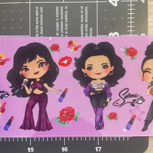 May include: Purple and black ribbon with a repeating pattern of cartoon images of Selena Gomez, roses, and lipstick. The ribbon has the text "Selena" written on it.