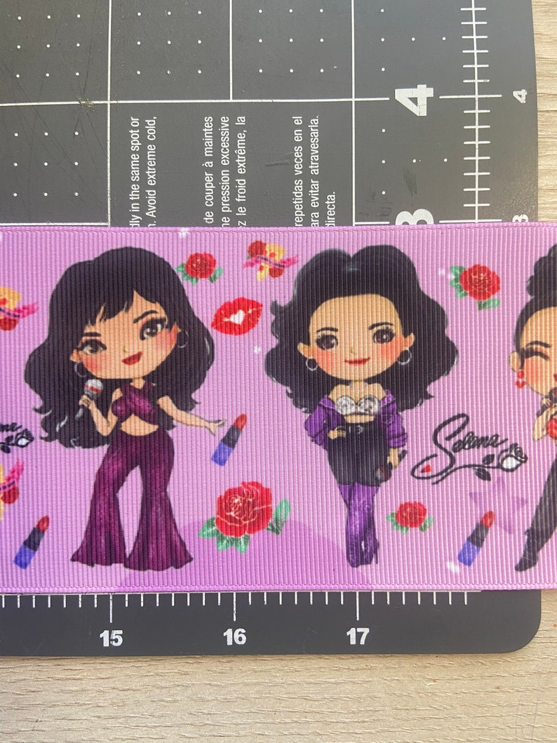 May include: Purple and black ribbon with a repeating pattern of cartoon images of Selena Gomez, roses, and lipstick. The ribbon has the text "Selena" written on it.