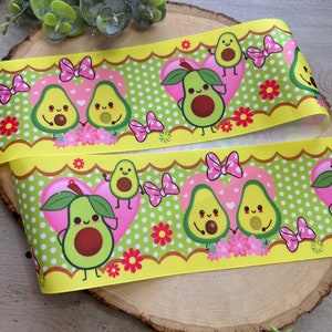 May include: Yellow and green ribbon with a repeating pattern of avocado halves with faces and pink bows. The avocados are holding hands and have hearts around them. The background is green with white polka dots and pink flowers.