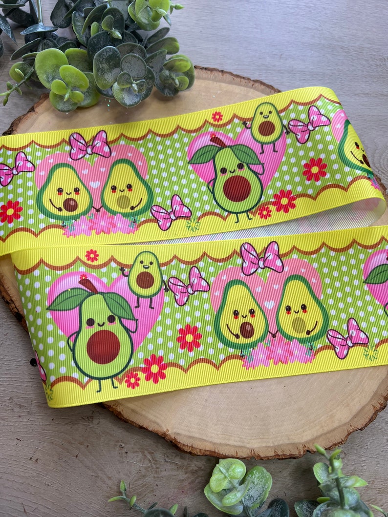 May include: Yellow and green ribbon with a repeating pattern of avocado halves with faces and pink bows. The avocados are holding hands and have hearts around them. The background is green with white polka dots and pink flowers.