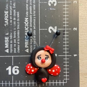May include: A black and red ladybug with white polka dots, a red heart, and a red bow. The ladybug is made of polymer clay and is approximately 2 cm in size.