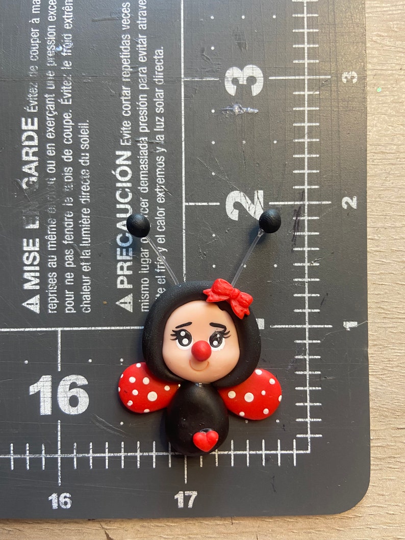 May include: A black and red ladybug with white polka dots, a red heart, and a red bow. The ladybug is made of polymer clay and is approximately 2 cm in size.