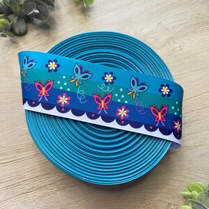 Pu&ograve; includere: Un rotolo di nastro blu turchese con un bordo bianco a festoni. Il nastro presenta un motivo ripetitivo di farfalle colorate e fiori bianchi.