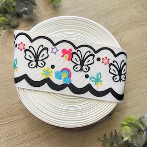 May include: A white ribbon with a black scalloped edge and a colorful print of butterflies, flowers, and a rainbow.