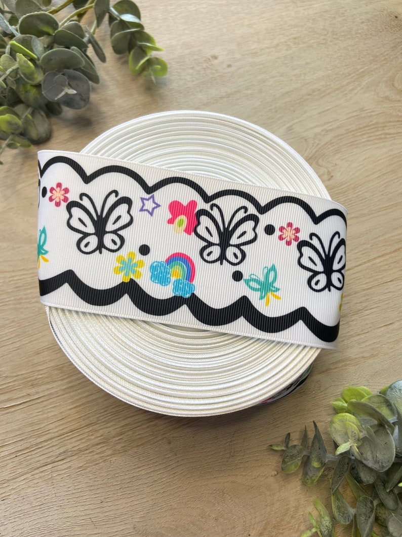 May include: A white ribbon with a black scalloped edge and a colorful print of butterflies, flowers, and a rainbow.