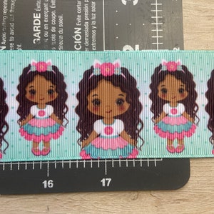 May include: A light blue ribbon with a repeating pattern of cartoon girls wearing pink and blue dresses and flower hair accessories. The ribbon has a polka dot background.