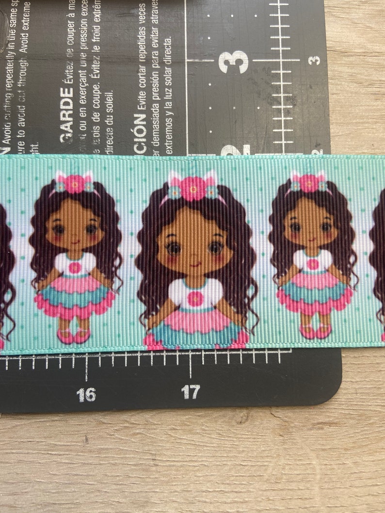 May include: A light blue ribbon with a repeating pattern of cartoon girls wearing pink and blue dresses and flower hair accessories. The ribbon has a polka dot background.