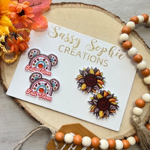 May include: A white canvas displays two pairs of decorative earrings. One pair features a rainbow design with a pumpkin and the text "Fall Vibes." The other pair showcases sunflower designs with leopard print centers. The canvas is on a wooden surface.