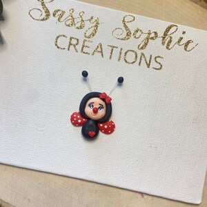 May include: A black and red ladybug with white polka dots and a red heart on its belly. The ladybug has a black face with white eyes and a red mouth. The ladybug is on a white canvas with the text "Sassy Sophie Creations" in gold glitter.