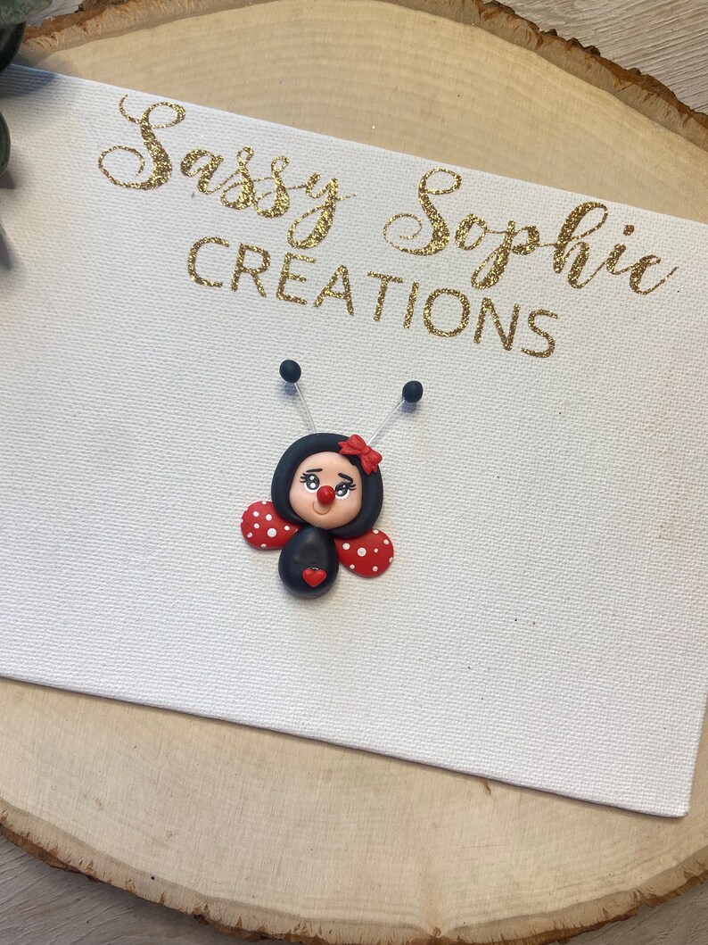 May include: A black and red ladybug with white polka dots and a red heart on its belly. The ladybug has a black face with white eyes and a red mouth. The ladybug is on a white canvas with the text "Sassy Sophie Creations" in gold glitter.