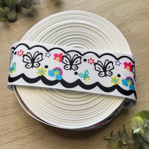 May include: A white ribbon with a black scalloped edge and a colorful butterfly, rainbow, and flower design.