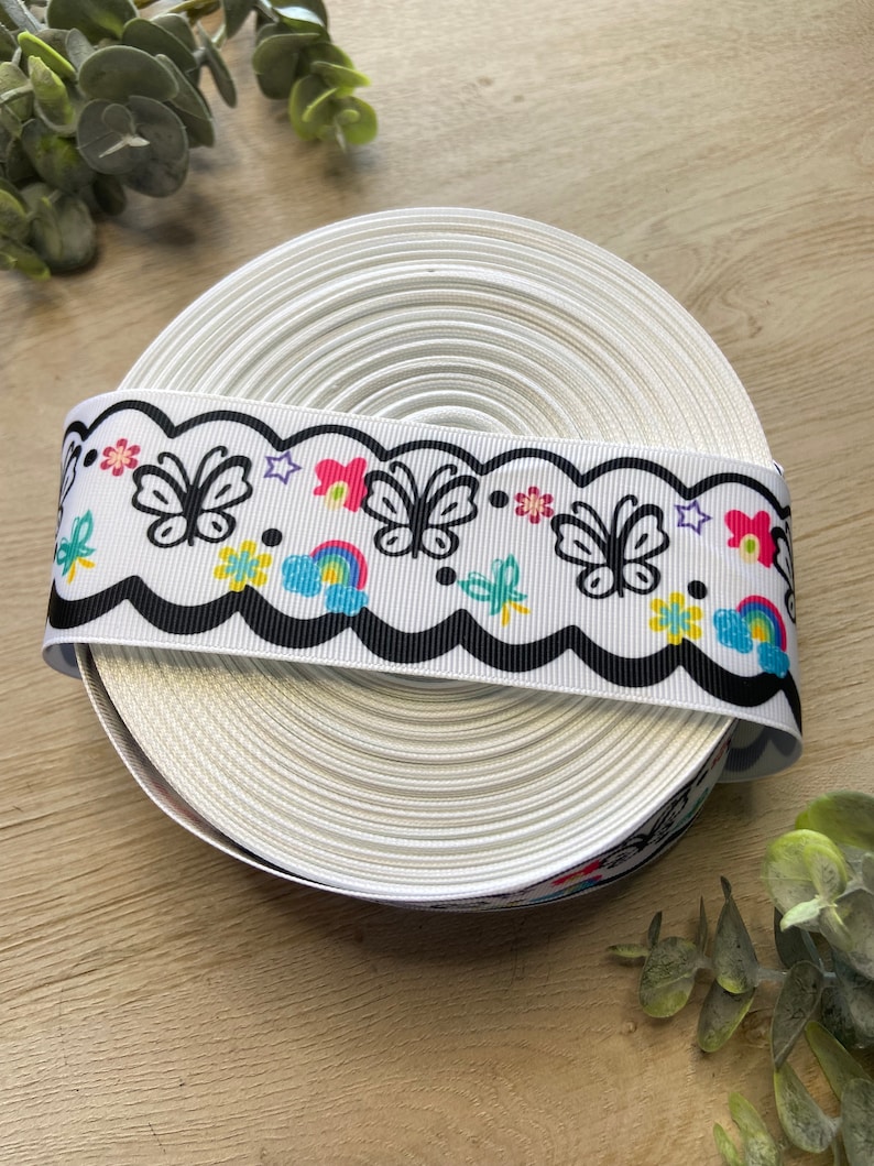 May include: A white ribbon with a black scalloped edge and a colorful butterfly, rainbow, and flower design.