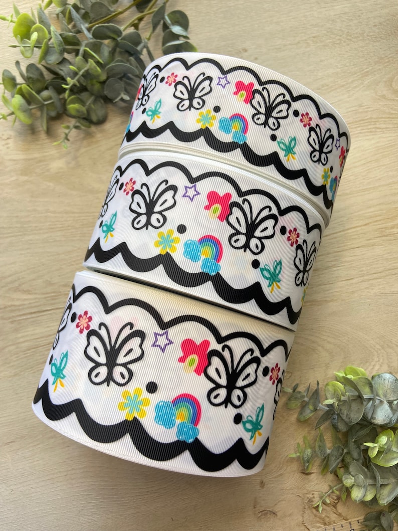 May include: Three rolls of white ribbon with a black scalloped edge. The ribbon is decorated with a colorful print of butterflies, flowers, rainbows, and stars.