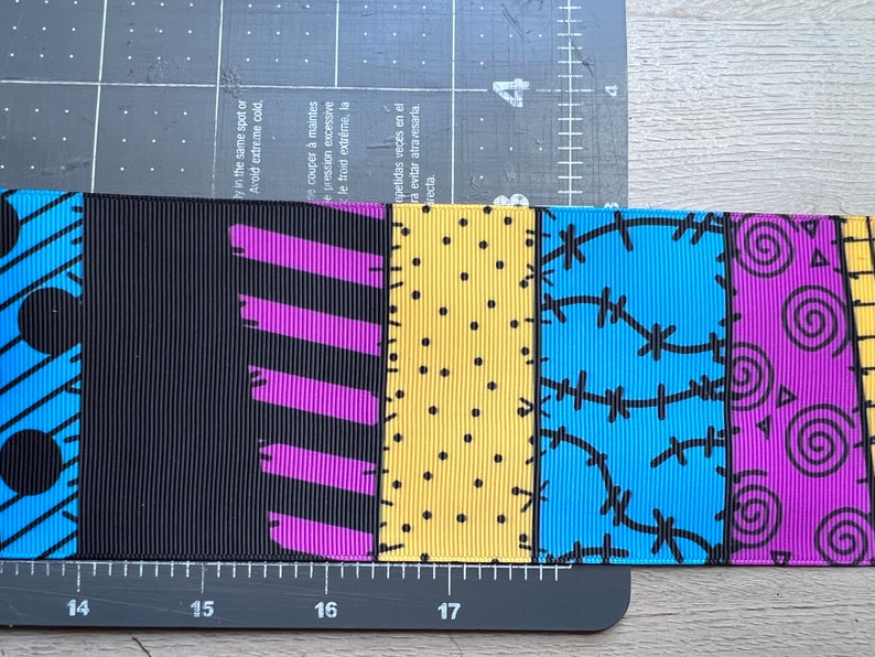 May include: Ribbon with a patchwork design featuring various patterns and colors. The ribbon includes black, purple, yellow, and blue sections with polka dots, stitches, and swirls. The ribbon is approximately 2 inches wide.