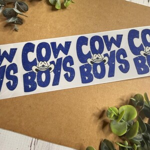 May include: White ribbon with the words "COWBOYS" and "COW" in blue, with a cowboy hat graphic. The ribbon is on a brown surface with green foliage.
