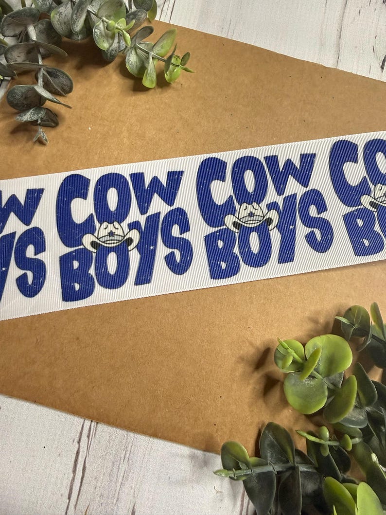 May include: White ribbon with the words "COWBOYS" and "COW" in blue, with a cowboy hat graphic. The ribbon is on a brown surface with green foliage.
