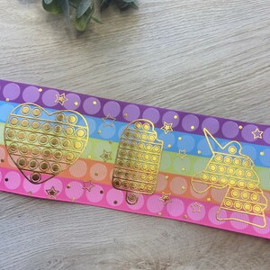 Pu&ograve; includere: Un nastro a righe arcobaleno con accenti dorati. Il nastro presenta un cuore, un ghiacciolo e un unicorno con un design pop-it.