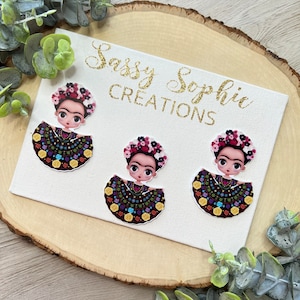 May include: Three colorful, hand-painted, round, resin,  Frida Kahlo-inspired ornaments with a black background, gold accents, and floral details. The ornaments are on a white canvas with the text "Sassy Sophia Creations" in gold glitter.